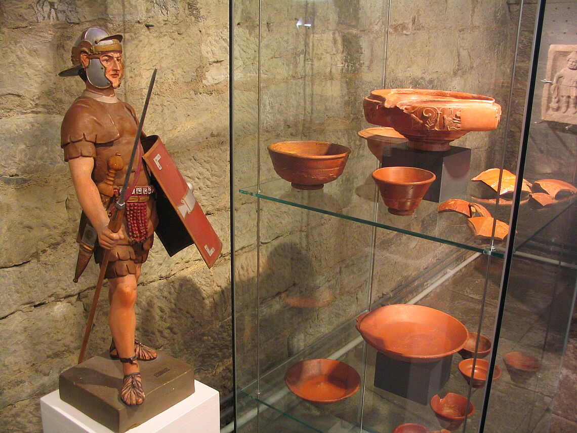Im Vordergrund ist die Figur eines römischen Soldaten zu sehen, im Hintergrund in einer Vitrine sieht man römsiches Geschirr.