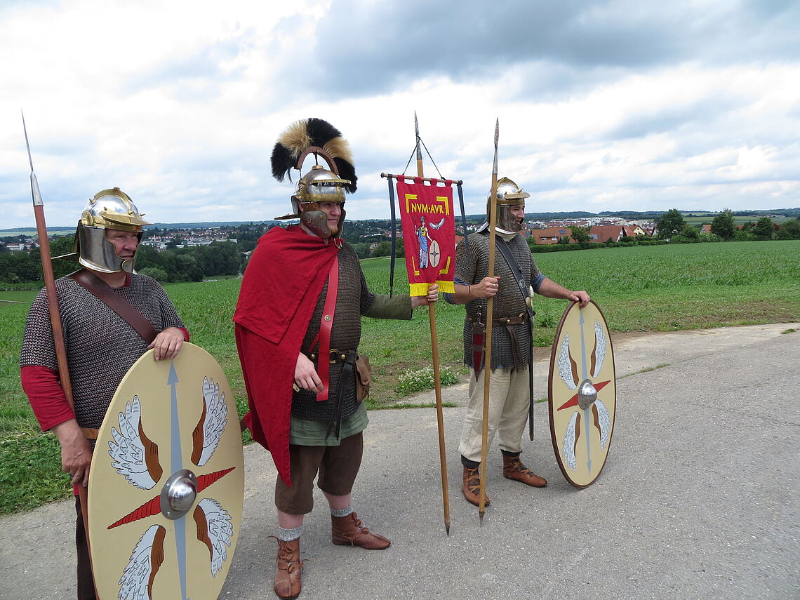 Drei Männer als Römersoldaten verkleidet sind zu sehen. Die äußeren haben jeweils ein Schild vor sich stehen.