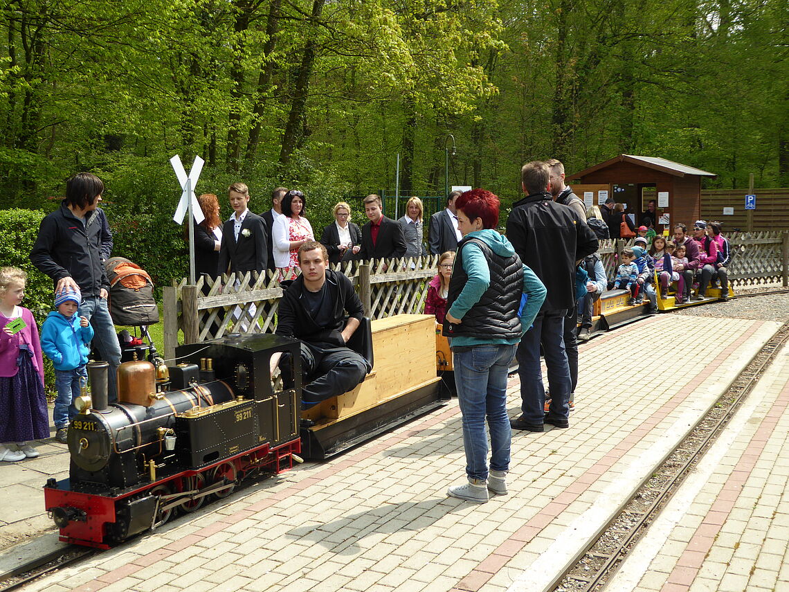 Eine Dampfgartenbahn fährt vorüber. Der Zugführer und weitere Passagiere sowie Zuschauer sind zu sehen.