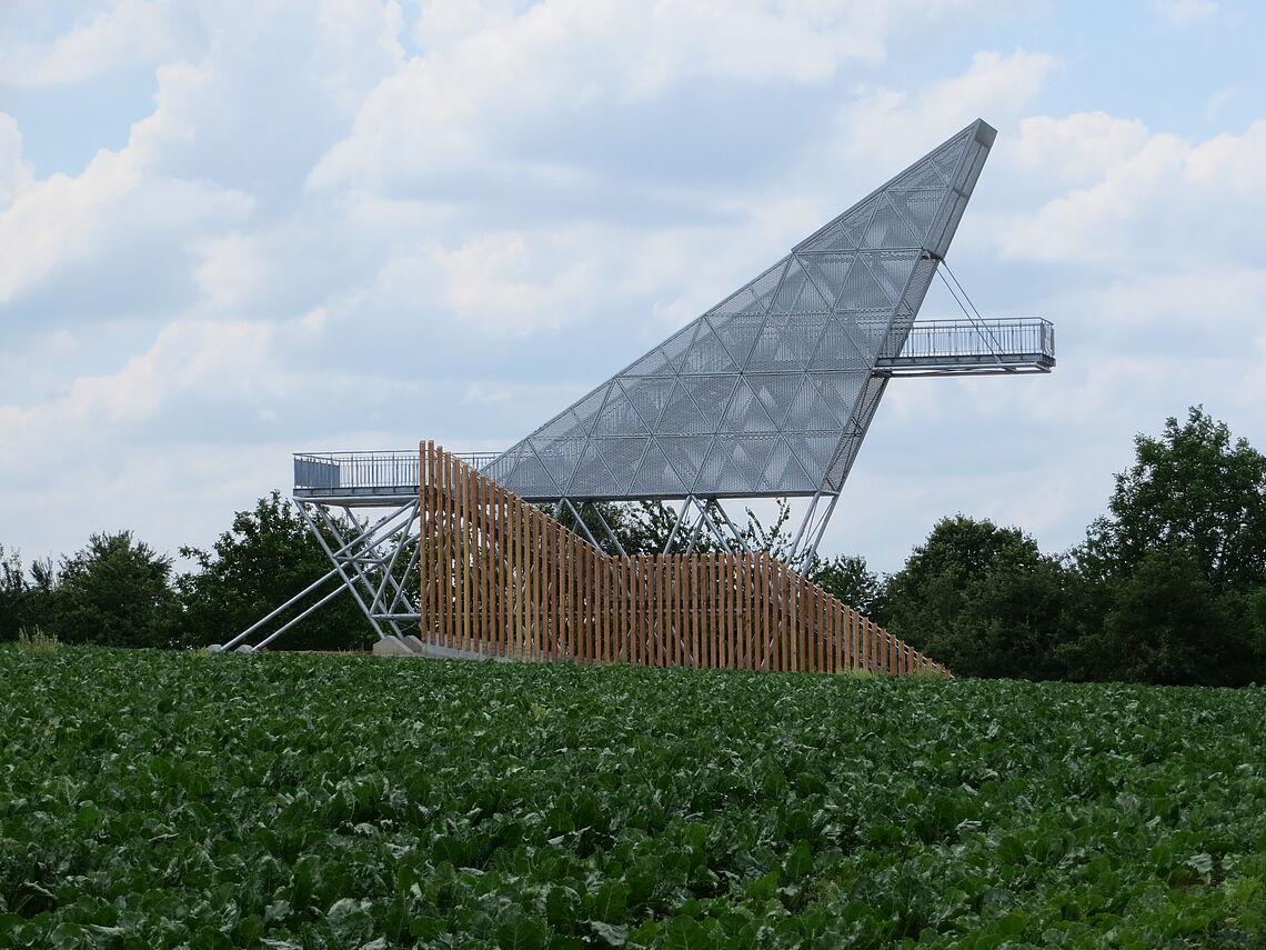 Die Aussichtsplattform Limes Blick in Öhringen ist zu sehen. Ein Konstruktion aus Stahl und Holz. Im Vordergrund ist ein grünes Feld zu sehen.