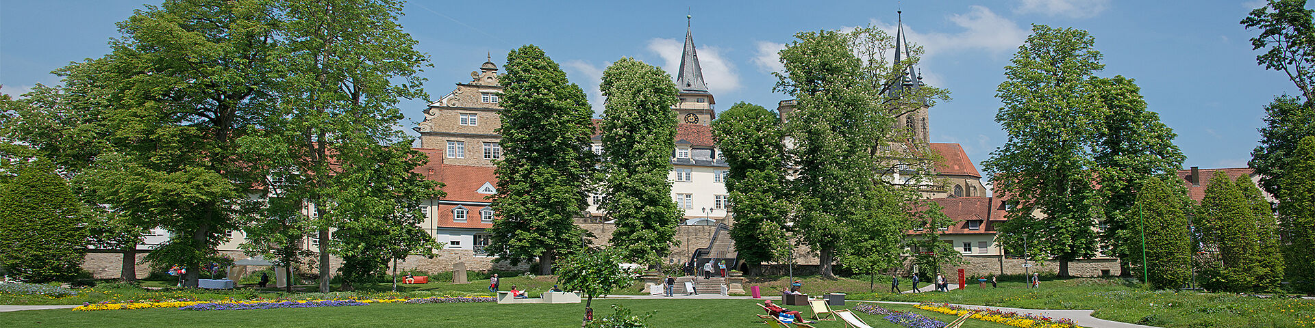 Der sommerliche Hofgarten in Öhringen ist zu sehen. Im Hintergrund hinter hohen Bäumen sieht man das Schloss und die Stiftskirche.