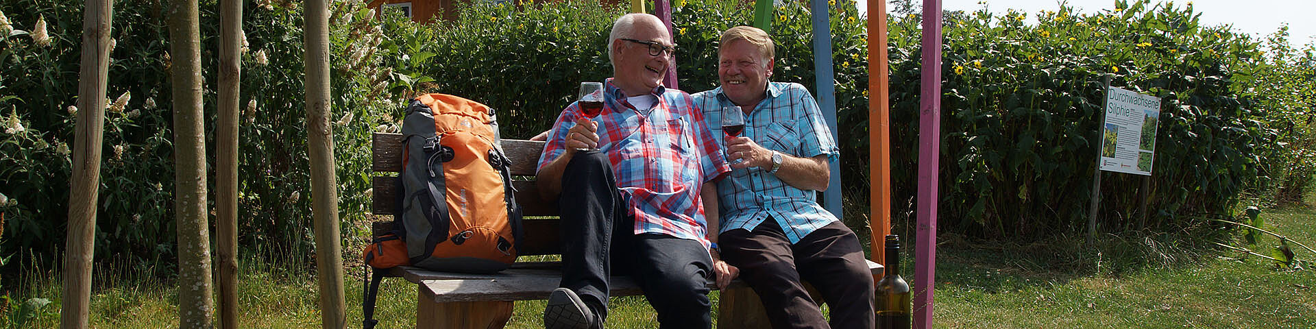 Zwei Männer sitzen mit einem Wanderrucksack und Weingläsern auf einer Bank. Im Hintergrund sind die bunten Stelen der Landesgartenschau Öhringen 2016 zu erkennen.